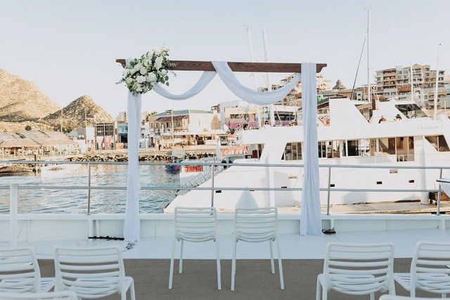 Ceremony arch draped in white fabric with rows of white chairs on a catamaran deck in the marina