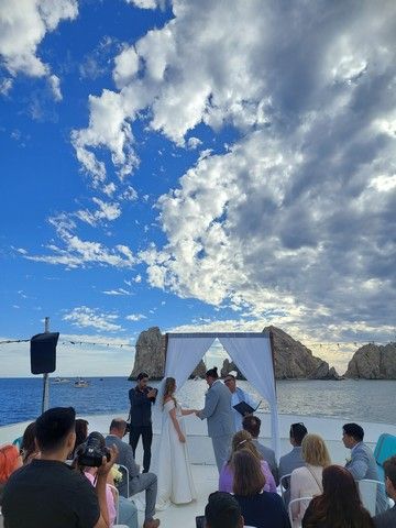 Wedding ceremony under a floral arch on a catamaran deck beneath a wide blue sky, with Land's End beyond